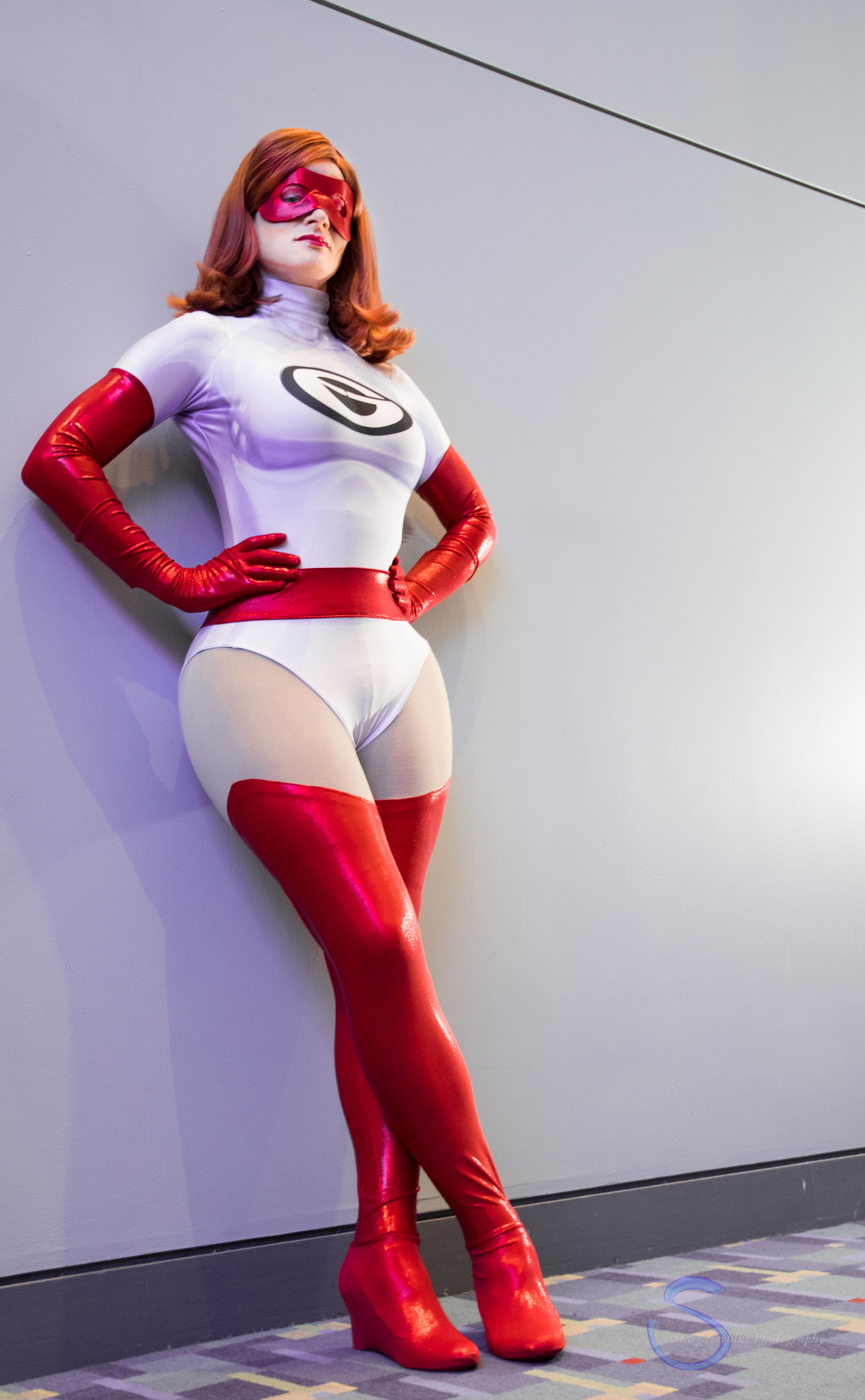 Jay is wearing a cosplay of Elastigirl's classic red and white costume from the Incredibles. She is leaning against a wall with her hands on her hips and her ankles crossed.
