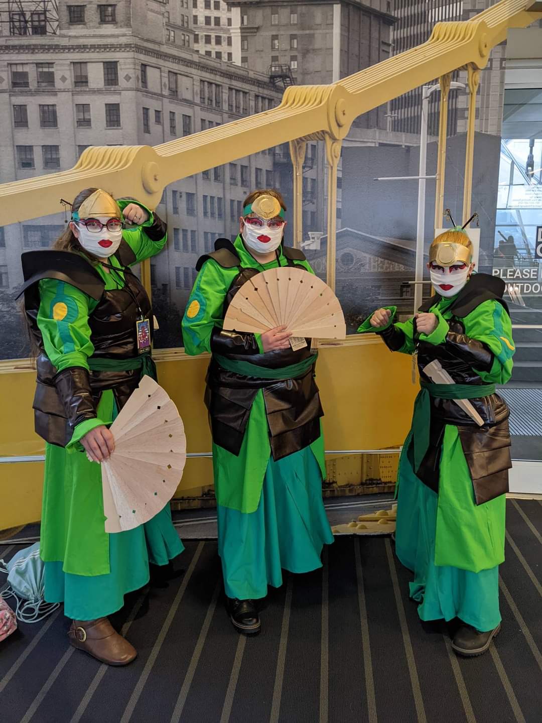 Three female cosplayers. They are dressed in dark green skirt with diagonal pleats, light green long jackets with long poofy sleeves, and leather (plether) armor and wrist guards. They have white face paint with red eye makeup. Due to covid had to wear white masks with red lips to stay safe but keep the look. They are holding large wooden fans.